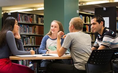 Students seated at a table, in discussion, with library books displayed on shelving behind them.