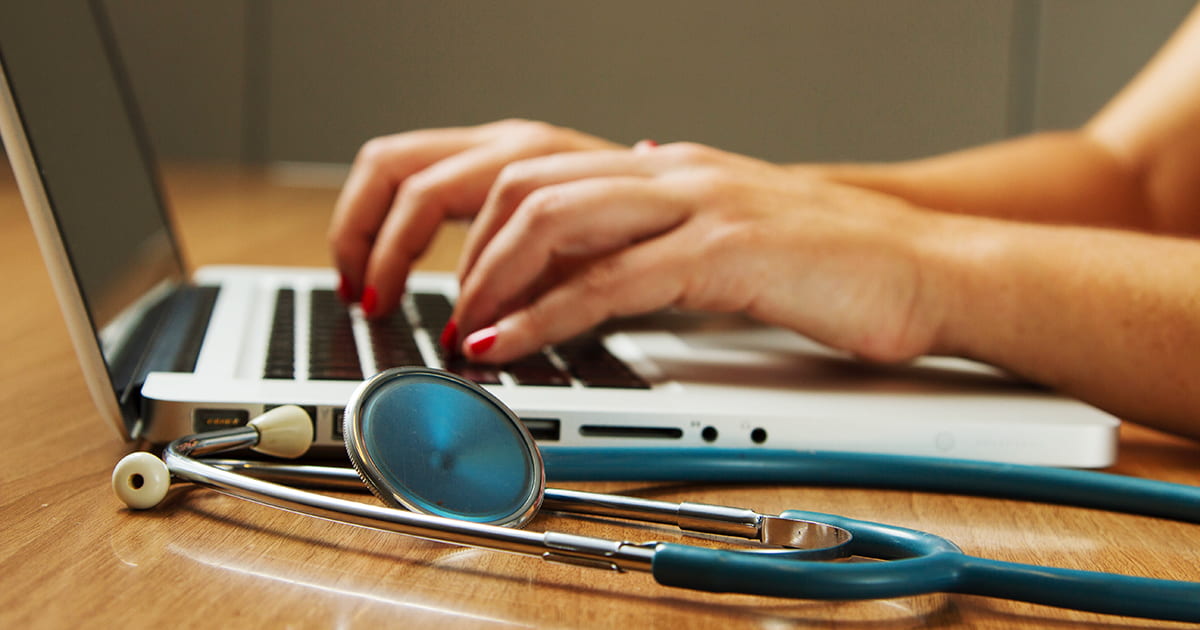 View of hands typing on a laptop keyboard with a stethoscope next to the laptop.